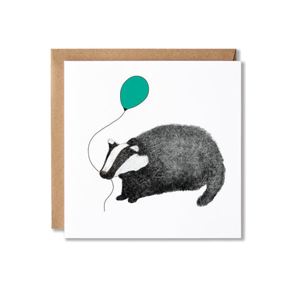 A square white greeting card with a brown kraft envelope behind it, featuring a black-and-white illustration of a badger holding a teal balloon.