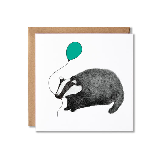A square white greeting card with a brown kraft envelope behind it, featuring a black-and-white illustration of a badger holding a teal balloon.