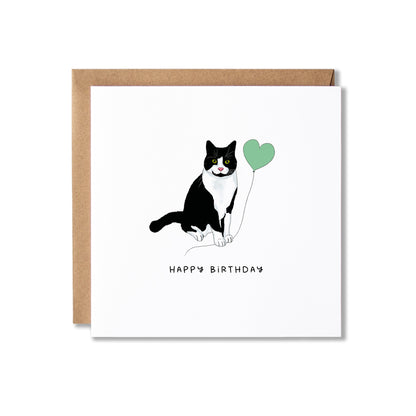 A square white birthday card with a brown kraft envelope behind it, featuring a black-and-white cat holding a mint green heart balloon and the words 'HAPPY BIRTHDAY' printed below.