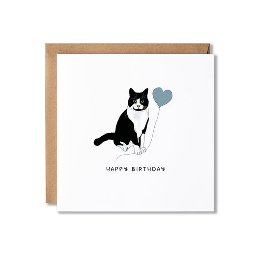 A square white birthday card with a kraft brown envelope behind it, featuring a black-and-white cat holding a blue heart balloon and the words 'HAPPY BIRTHDAY' below.