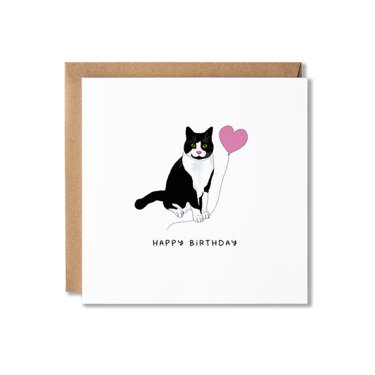 A square white birthday card with a brown kraft envelope behind it, featuring a black-and-white cat holding a pink heart balloon and the words 'HAPPY BIRTHDAY' below.