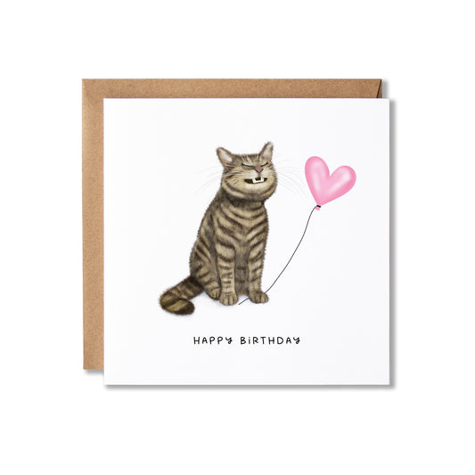 A square birthday card with a brown kraft envelope, featuring an illustration of a grinning tabby cat sitting next to a pink heart-shaped balloon and the text 'HAPPY BIRTHDAY' below it.