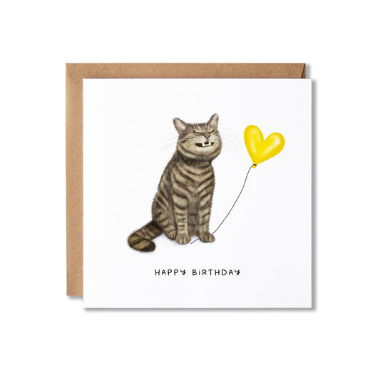 A square birthday card with a brown kraft envelope, featuring an illustration of a grinning grey tabby cat sitting next to a yellow heart-shaped balloon and the text 'HAPPY BIRTHDAY' below.