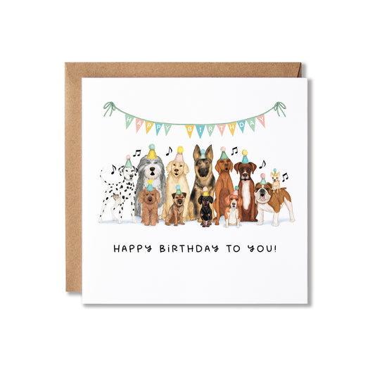 A square white birthday card with a kraft brown envelope behind it, featuring an illustration of a group of dogs wearing party hats under a colorful banner, with the text 'HAPPY BIRTHDAY TO YOU!' printed below.