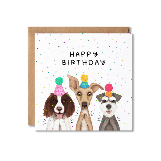 A square birthday card with a kraft envelope behind it, featuring three illustrated dogs wearing party hats under the words 'HAPPY BIRTHDAY' on a white background with scattered confetti.