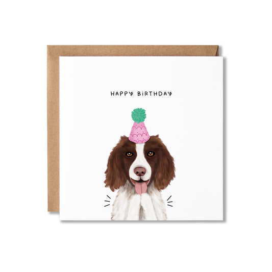A square white birthday card with a brown envelope behind it, featuring an illustration of a brown and white dog wearing a pink party hat with a green pom-pom. The text 'HAPPY BIRTHDAY' appears above the dog.
