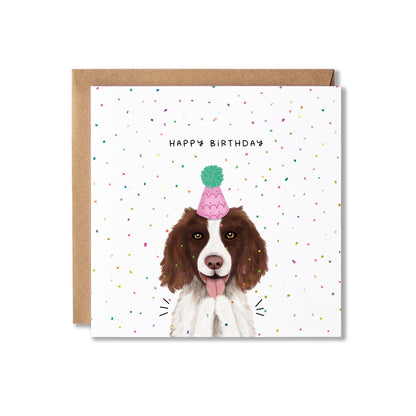 A square birthday card with a brown envelope behind it, featuring an illustration of a brown and white dog wearing a pink party hat with a green pom-pom. The card is decorated with small multicolored confetti and reads 'HAPPY BIRTHDAY'.