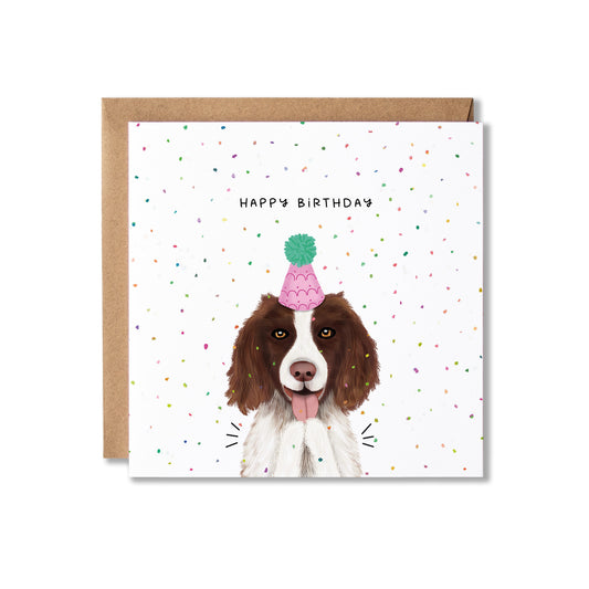 A square birthday card with a brown envelope behind it, featuring an illustration of a brown and white dog wearing a pink party hat with a green pom-pom. The card is decorated with small multicolored confetti and reads 'HAPPY BIRTHDAY'.