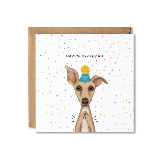 A square birthday card with a brown kraft envelope behind it. The card features a white background scattered with small multicolored confetti and the words 'HAPPY BIRTHDAY' above an illustration of a light brown dog wearing a blue party hat with a yellow pom-pom.
