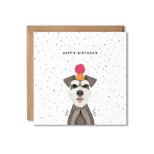 A square birthday card with a brown kraft envelope behind it. The card features a white background with scattered multicolored confetti and the text 'HAPPY BIRTHDAY' above an illustrated gray and white dog wearing a party hat with a pink pom-pom.
