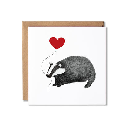 A square white greeting card with a kraft brown envelope behind it, featuring a black-and-white illustration of a badger holding a red heart-shaped balloon.