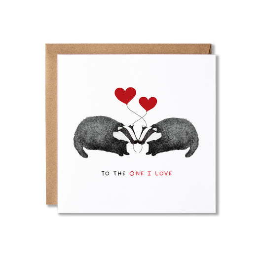 A square white greeting card with a kraft brown envelope behind it, featuring an illustration of two badgers facing each other with two red hearts above them and the text 'TO THE ONE I LOVE' below.