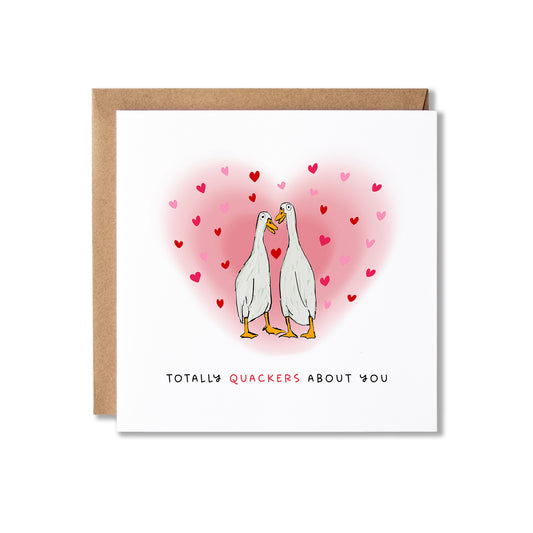 A square white greeting card with a brown kraft envelope behind it, featuring an illustration of two white quackers facing each other inside a pink heart made of small red hearts. The text at the bottom reads 'TOTALLY QUACKERS ABOUT YOU'.