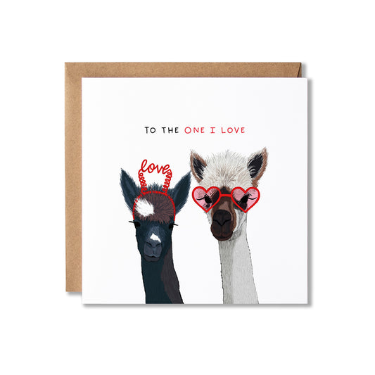 A square greeting card with a kraft envelope behind it, featuring an illustration of two alpacas wearing red heart-shaped glasses and red headband with the word 'love' written above them. The card reads 'TO THE ONE I LOVE' at the top.