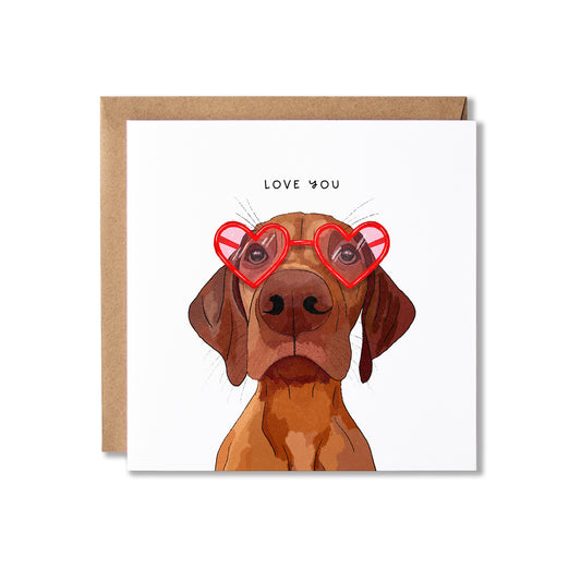 A square greeting card with a brown envelope behind it, featuring an illustration of a Vizsla dog wearing red heart-shaped glasses and the words 'LOVE YOU' above it, on a white background.