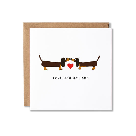 A white greeting card with a brown kraft envelope behind it, featuring a cartoon dachshund facing another dachshund with a small red heart between them and the text 'LOVE YOU SAUSAGE' below.