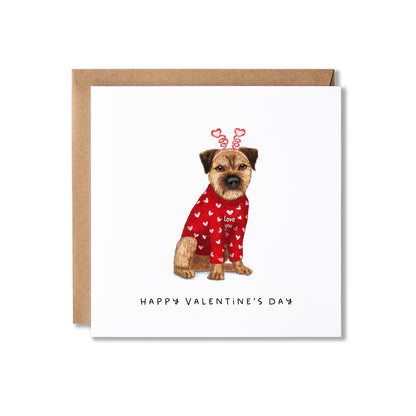 A square Valentine's Day greeting card with a brown kraft envelope behind it. The card features an illustration of a border terrier dog wearing a red sweater covered in white hearts, with red heart-shaped antlers, and the text 'HAPPY VALENTINE'S DAY' printed below.