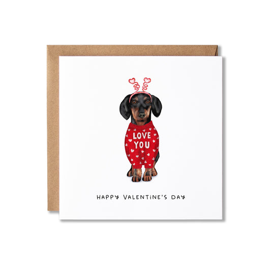 A square Valentine's Day greeting card with a kraft envelope behind it. The card features an illustration of a dachshund wearing a red sweater with white hearts and the words 'LOVE YOU', with 'HAPPY VALENTINE'S DAY' printed below.