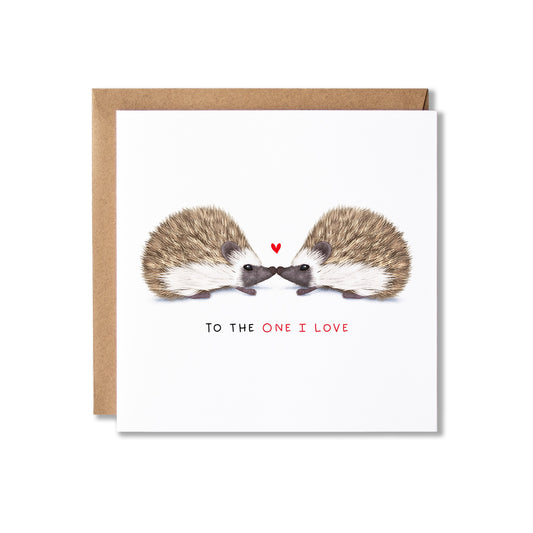 A square greeting card with a brown kraft envelope behind it, featuring an illustration of two hedgehogs facing each other with a small red heart above them and the text 'TO THE ONE I LOVE' below.
