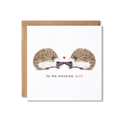 A square greeting card with a brown envelope behind it, featuring an illustration of two hedgehogs facing each other with a small red heart between them and the text 'TO MY AMAZING WIFE' below.