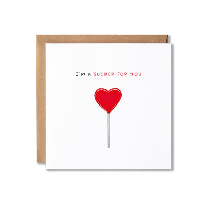 A square white greeting card with a brown kraft envelope behind it, featuring the text 'I'M A SUCKER FOR YOU' above a red heart-shaped lollipop illustration on a white background.