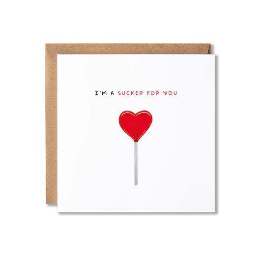A square white greeting card with a brown kraft envelope behind it, featuring the text 'I'M A SUCKER FOR YOU' above a red heart-shaped lollipop illustration on a white background.