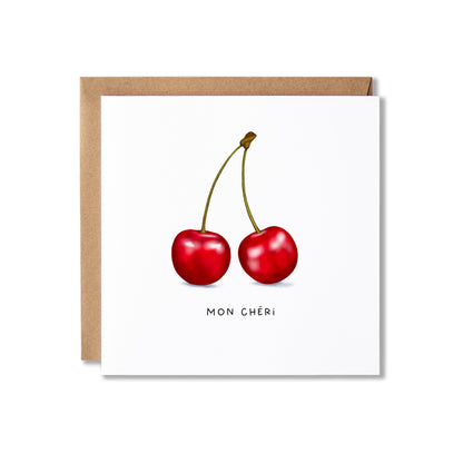 A square white greeting card with an illustration of two red cherries and the text 'MON CHÉRI' below, shown with a brown kraft envelope behind it on a white background.