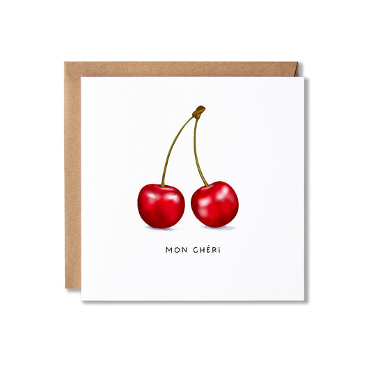 A square white greeting card with an illustration of two red cherries and the text 'MON CHÉRI' below, shown with a brown kraft envelope behind it on a white background.