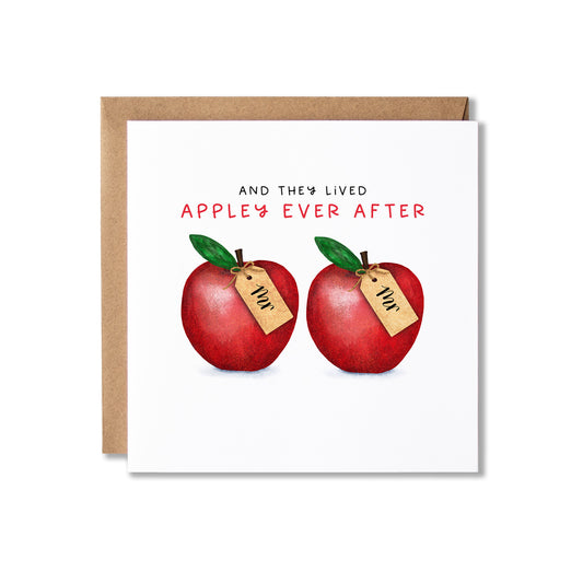 A square greeting card with a kraft envelope behind it, featuring the text 'AND THEY LIVED APPLE'S EVER AFTER' above two illustrated red apples with green leaves each with a small paper tag reading 'Mr'.