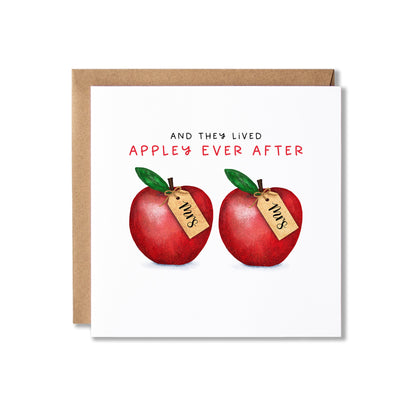 A square greeting card with a kraft envelope behind it, featuring the text 'AND THEY LIVED APPLE'S EVER AFTER' above two red apples with green leaves and small tags reading 'Mrs'.