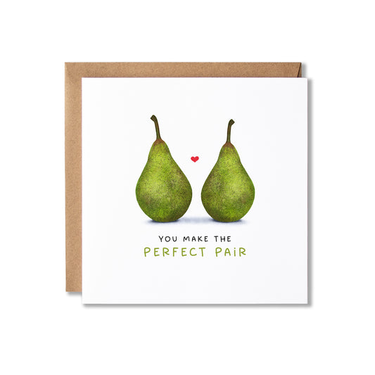 A square greeting card with a white front featuring an illustration of two green pears and a small red heart, with the text 'YOU MAKE THE PERFECT PAIR' below. A brown kraft envelope is partially visible behind the card on a white background.