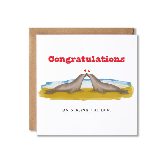 A square greeting card with a kraft envelope behind it, featuring an illustration of two seals on a beach with the text 'Congratulations ON SEALING THE DEAL'.