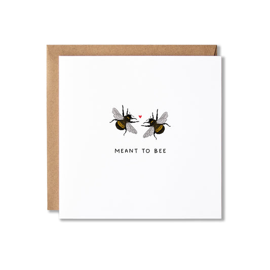A square white greeting card with a kraft brown envelope behind it, featuring an illustration of two bumblebees facing each other with a small red heart between them and the text 'MEANT TO BEE' below.