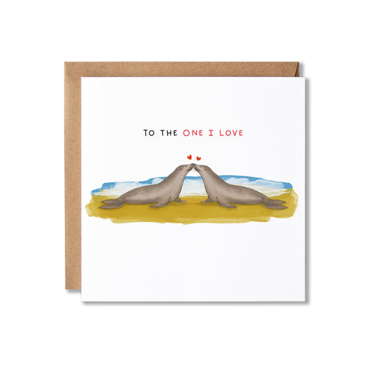 A square greeting card with a brown kraft envelope, featuring an illustration of two seals facing each other on a yellow beach with blue water and the text 'TO THE ONE I LOVE' above them.