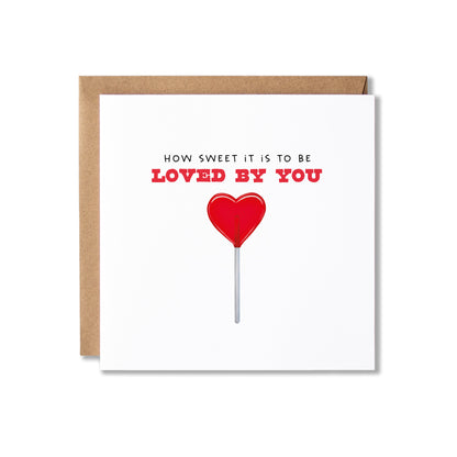 A square white greeting card with a kraft brown envelope behind it, featuring the text 'HOW SWEET IT IS TO BE LOVED BY YOU' in black and red above a red heart-shaped lollipop illustration.