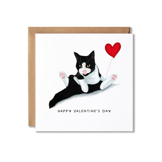 A square Valentine's Day greeting card with a brown kraft envelope behind it. The card features an illustration of a black-and-white cat lying down and holding a red heart-shaped balloon, with the text 'HAPPY VALENTINE'S DAY' printed below.