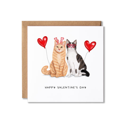 A square Valentine's Day greeting card with a kraft envelope behind it. The card features an illustration of two cats sitting side by side, one orange tabby and one black-and-white cat, each holding a red heart-shaped balloon with Happy Valentine's Day written underneath.