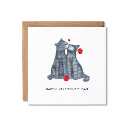 A square Valentine's Day greeting card with a white front featuring an illustration of two grey cats facing each other with red flowers and a small red heart above them with Happy Valentine's Day written underneath. The card is shown with a brown kraft envelope behind it on a white background.