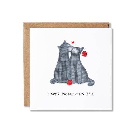 A square Valentine's Day greeting card with a white front featuring an illustration of two grey cats facing each other with red flowers and a small red heart above them with Happy Valentine's Day written underneath. The card is shown with a brown kraft envelope behind it on a white background.