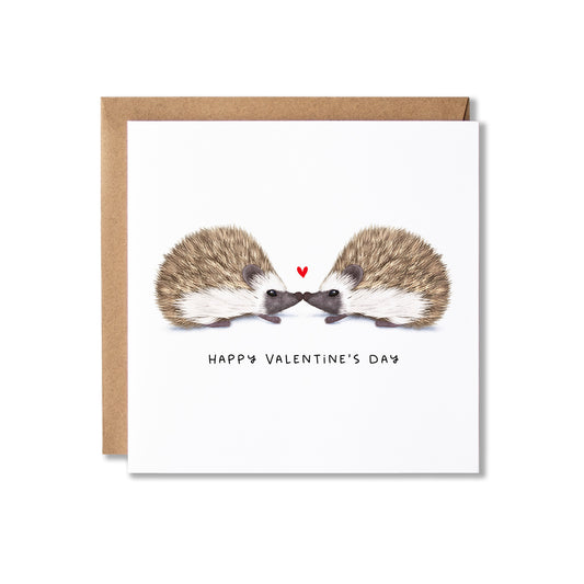 A square Valentine's Day greeting card with a brown kraft envelope behind it. The card features an illustration of two hedgehogs facing each other with a small red heart between them and the text 'HAPPY VALENTINE'S DAY' below.
