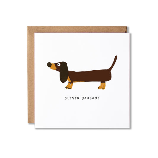 A square white greeting card with a brown dachshund illustration and the text 'CLEVER SAUSAGE' underneath, shown with a brown kraft envelope behind it on a light gray background.
