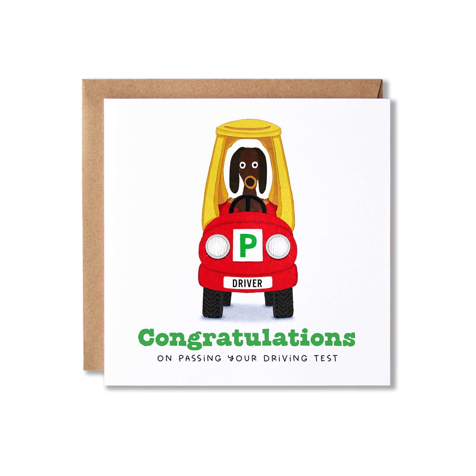 A square white greeting card with a brown kraft envelope behind it, featuring a cartoon dog driving a red car with a green 'P' plate and the text 'Congratulations on passing your driving test.'.