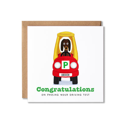 A square white greeting card with a brown kraft envelope behind it, featuring a cartoon dog driving a red car with a green 'P' plate and the text 'Congratulations on passing your driving test.'.