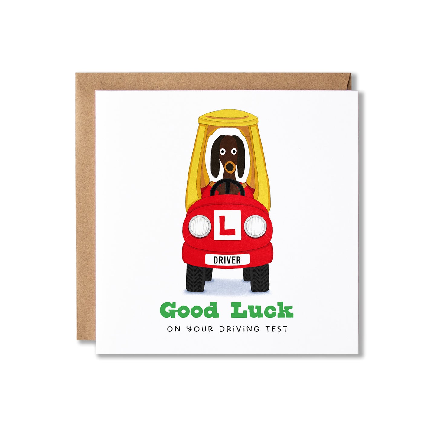 A square greeting card with a brown kraft envelope behind it, featuring a cartoon dog driving an red car with an L-shaped plate and the text 'Good Luck ON YOUR DRIVING TEST'.