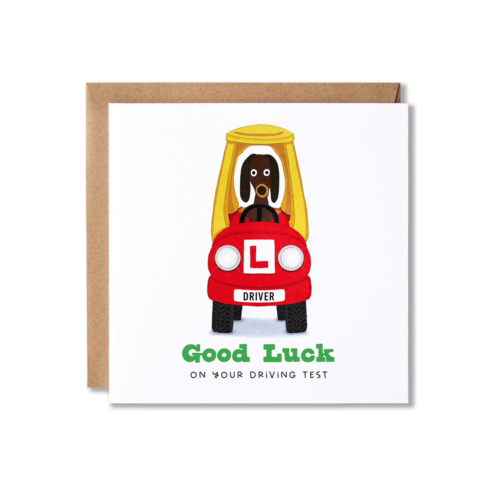 A square greeting card with a brown kraft envelope behind it, featuring a cartoon dog driving an red car with an L-shaped plate and the text 'Good Luck ON YOUR DRIVING TEST'.