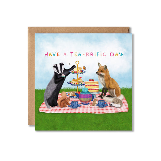 A square greeting card with a brown kraft envelope behind it, featuring an illustrated Afternoon tea picnic scene with a badger, fox, rabbit, hedgehog, cakes, teapot, and the text 'HAVE A TEA-RRIFIC DAY' on a grassy background.