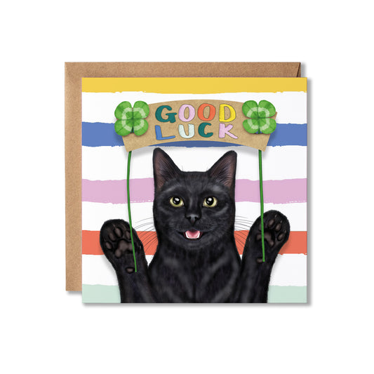 A square greeting card with a kraft envelope behind it, featuring a black cat behind a sign that reads “GOOD LUCK” with green four-leaf clovers and pastel horizontal stripes.