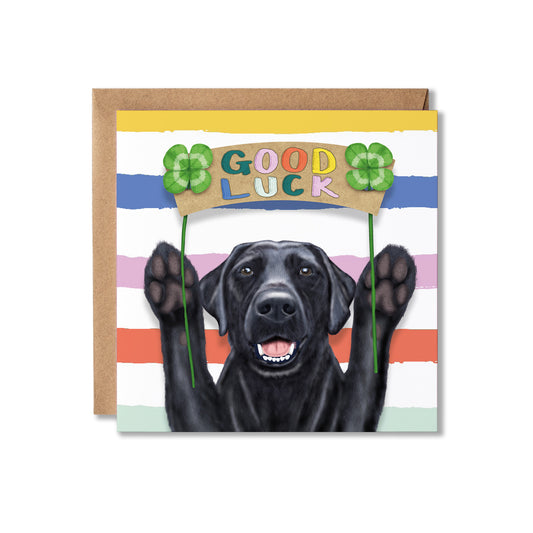 A square greeting card with a kraft envelope behind it, featuring a black dog holding a banner that says GOOD LUCK with four-leaf clovers on a striped background.