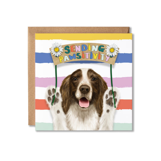 A square greeting card with a brown envelope behind it, featuring a Spaniel dog illustration holding a sign that reads 'SENDING PAWSITIVITY' against pastel horizontal stripes.