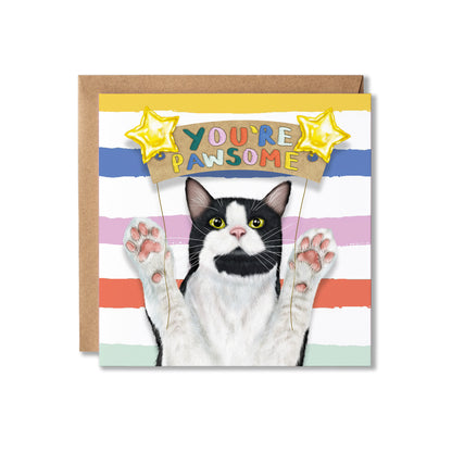 A square greeting card featuring a black-and-white cat with paws raised, holding a banner that reads 'YOU'RE PAWSOME,' set against pastel horizontal stripes. A brown kraft envelope is placed behind the card on a white background.